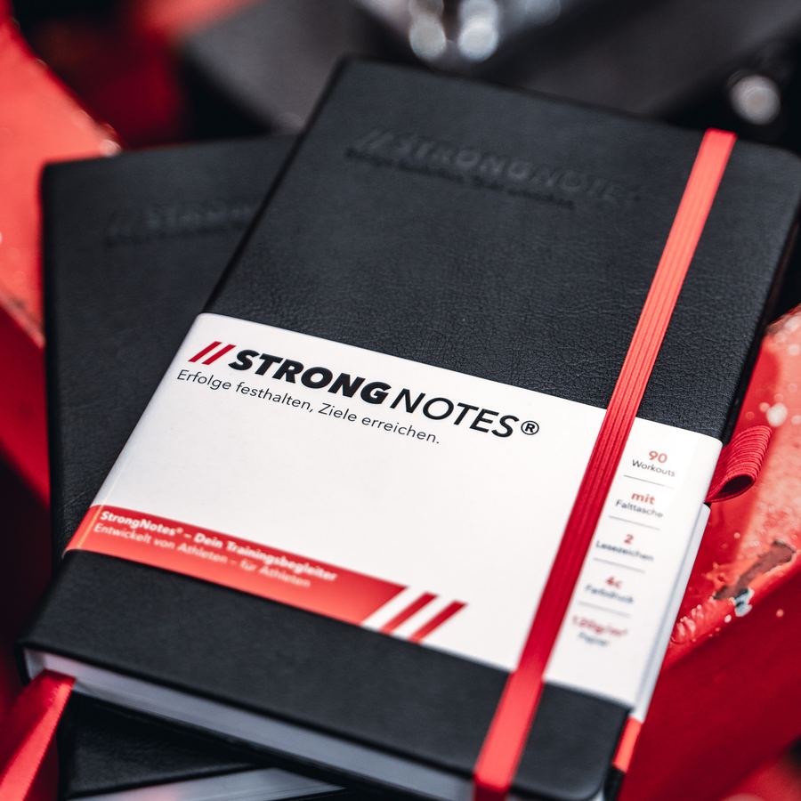 Zwei StrongNotes® Trainingsbücher mit Banderole und rotem Gummiband auf einer Hantelscheibe.