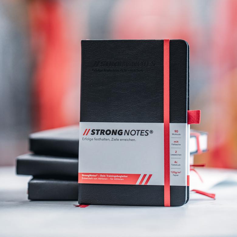 StrongNotes® Trainingsbuch mit Banderole und rotem Gummiband, dahinter ein Stapel weiterer Bücher.